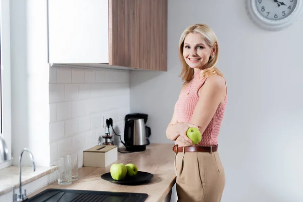 Mujer madura sosteniendo manzana y mirando a la cámara en la cocina - foto de stock