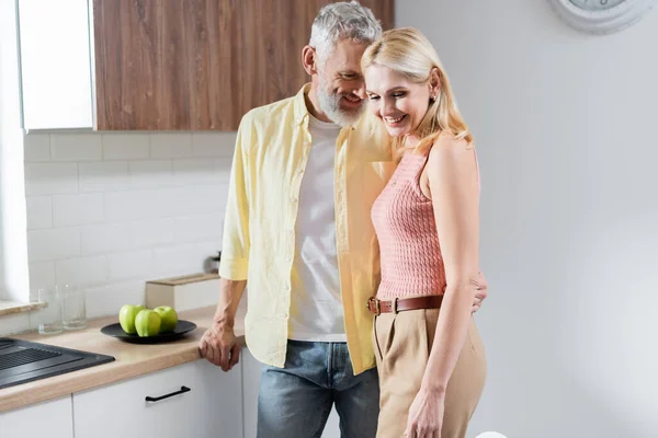 Smiling mature man hugging wife near apples on kitchen worktop — Stock Photo