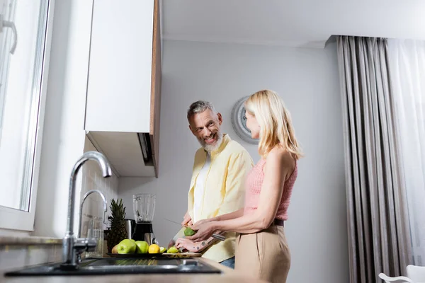 Homem positivo cortando frutas e olhando para a esposa na cozinha — Fotografia de Stock