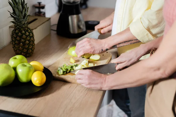 Ausgeschnittene Ansicht eines Paares mittleren Alters, das in der Küche Früchte in der Nähe eines verschwommenen Mixers schneidet — Stockfoto