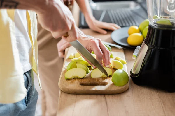 Cropped view of man cutting apple near blender and blurred wife in kitchen — Stock Photo