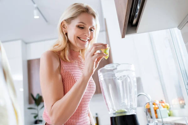 Mulher madura alegre derramando frutas no liquidificador na cozinha — Fotografia de Stock