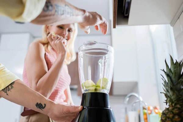Tattooed man holding blender with fruits near blurred wife in kitchen — Stock Photo