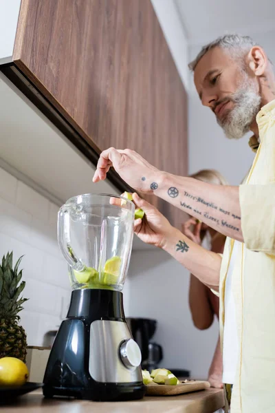 Vista a basso angolo dell'uomo tatuato che versa frutta nel frullatore in cucina — Foto stock