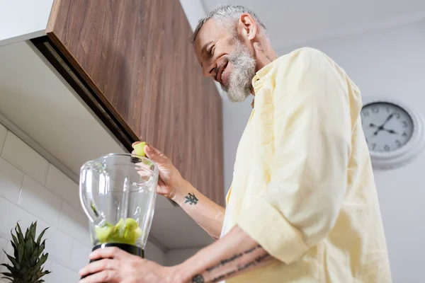 Low angle view of positive tattooed man pouring fruits in blender in kitchen — Stock Photo