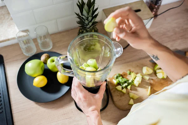 Cropped view of mature man holding apple and blender near blurred fruits in kitchen — Stock Photo