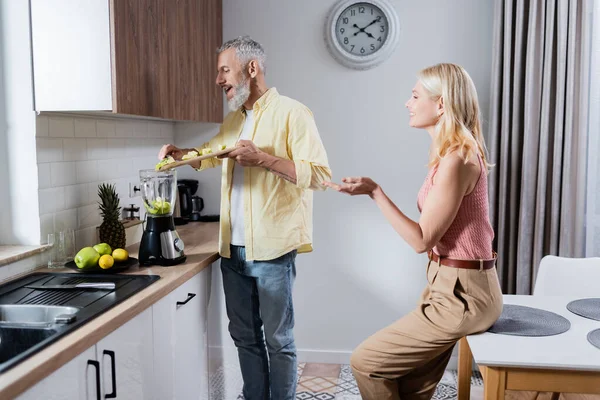 Side view of smiling woman sitting near husband preparing smoothie in kitchen — Stock Photo