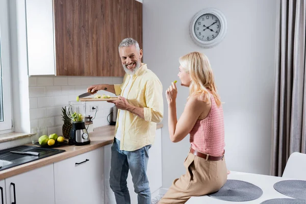 Hombre positivo añadiendo frutas en licuadora cerca de la esposa en la cocina - foto de stock