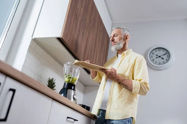 Vista de ángulo bajo del hombre tatuado que sostiene la tabla de cortar cerca de la licuadora con frutas en la cocina - foto de stock