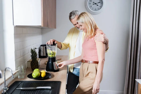 Sorrindo homem preparando smoothie e abraçando esposa na cozinha — Fotografia de Stock