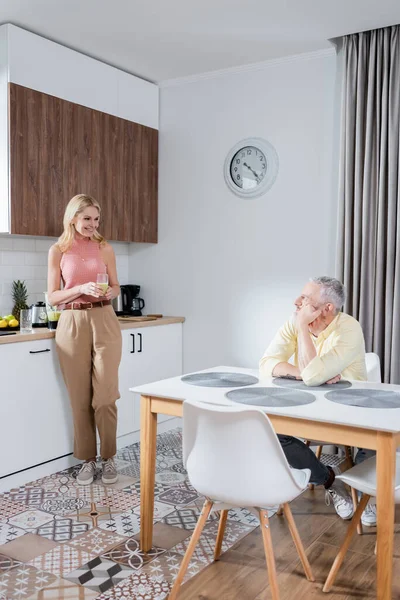 Cheerful mature woman holding fruit smoothie near husband in kitchen — Stock Photo