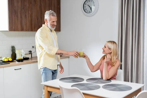 Sorrindo casal maduro clinking com copos de smoothie na cozinha — Fotografia de Stock