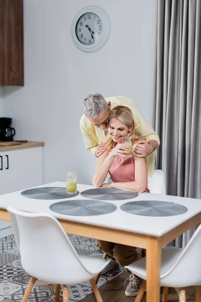Reifer Mann umarmt lächelnde Frau mit Glas Smoothie in Küche — Stockfoto