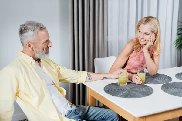 Позитивный зрелый мужчина смотрит на жену со стаканом смузи на кухне — стоковое фото