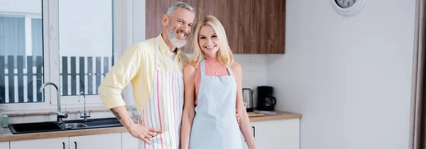 Pareja madura positiva en delantales mirando a la cámara en la cocina, pancarta - foto de stock