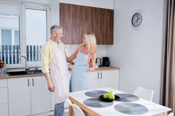 Side view of cheerful mature man in apron hugging blonde wife in kitchen — Stock Photo