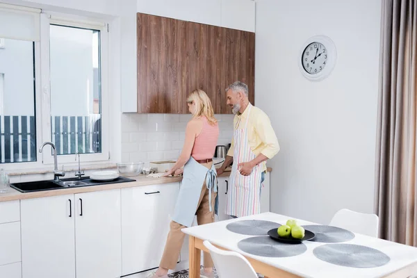 Усміхнений чоловік в фартусі стоїть біля дружини готує тісто на кухні — стокове фото