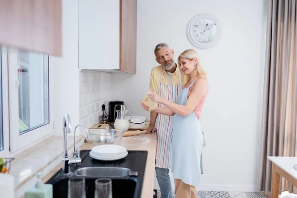 Зрелый мужчина, стоящий рядом с улыбающейся женой с баблом на кухне — стоковое фото