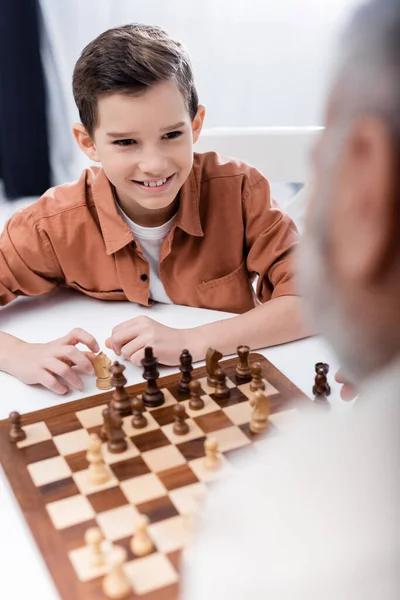 Cheerful boy playing chess with blurred grandpa at home — Stock Photo