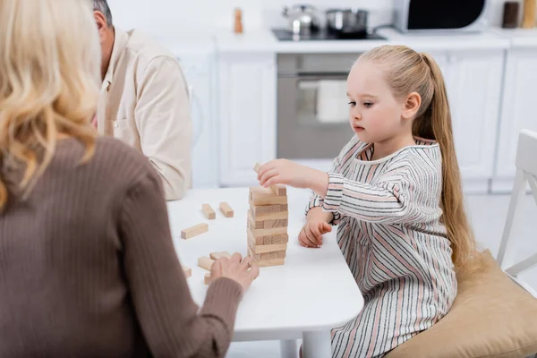 Menina jogando blocos de madeira jogo perto de avós borrados na cozinha — Fotografia de Stock