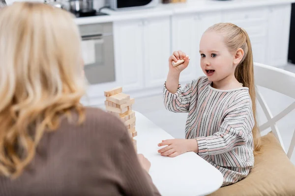 Chica sorprendida jugando bloques de madera juego cerca de la abuela borrosa en la cocina - foto de stock