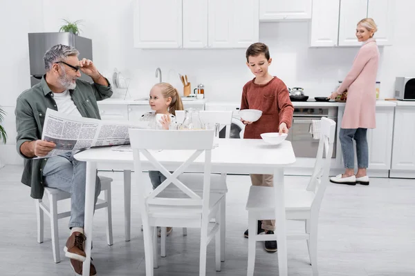 Uomo sorridente con giornale guardando nipotini apparecchiare la tavola per la colazione — Foto stock