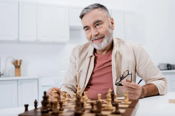 Pleased grey-haired man holding eyeglasses while playing chess in kitchen — Stock Photo