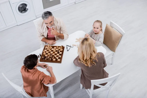 Высокий угол обзора девочки, смотрящей на камеру рядом с бабушкой и братом, играющими в шахматы на кухне — стоковое фото