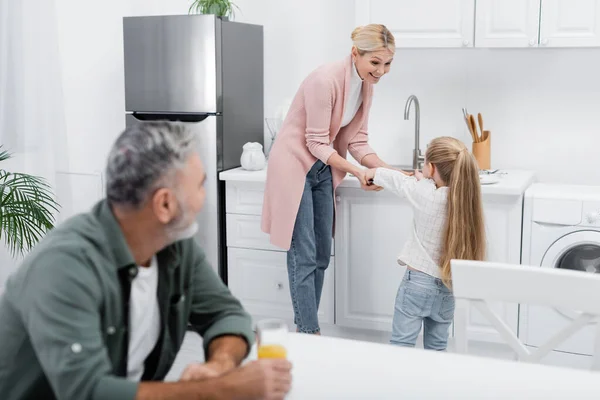 Senior man with orange juice looking at granddaughter helping granny in kitchen — Stock Photo