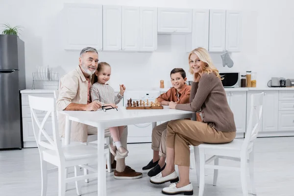 Feliz pareja con nietos sonriendo a la cámara mientras juega al ajedrez en la cocina - foto de stock