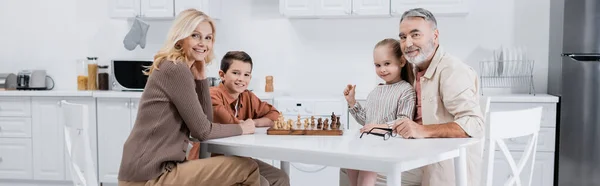 Счастливые дети с бабушкой и дедушкой смотрят в камеру во время игры в шахматы на кухне, баннер — стоковое фото