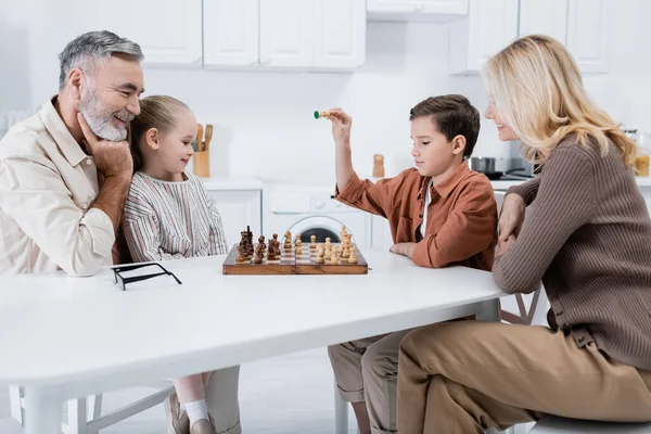 Ragazzo che tiene la figura degli scacchi vicino sorridente nonni e sorella in cucina — Foto stock