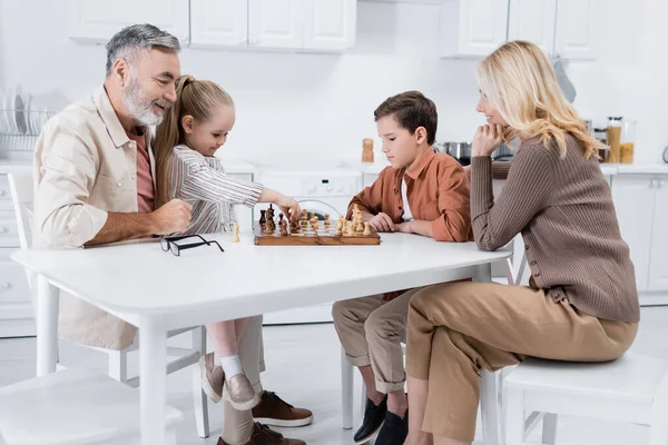 Niña sosteniendo la figura mientras juega al ajedrez con los abuelos y el hermano en la cocina - foto de stock