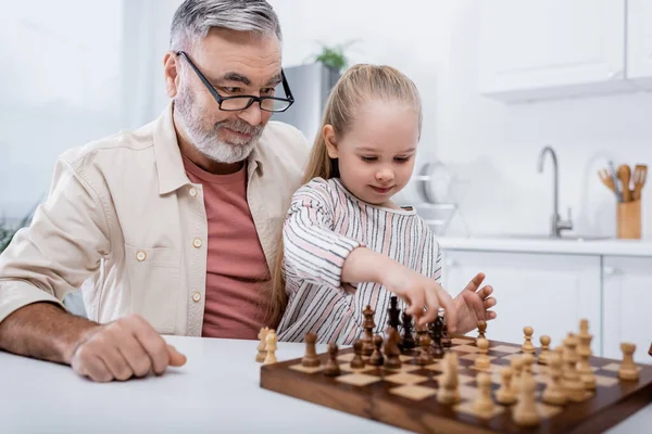 Senior man in eyeglasses smiling near granddaughter playing chess — Stock Photo