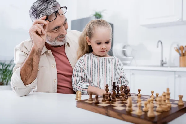 Happy senior man holding eyeglasses while playing chess with little granddaughter in kitchen — Stock Photo