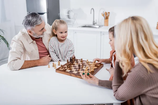 Femme floue tenant des chiffres près de mari et petits-enfants jouant aux échecs dans la cuisine — Photo de stock