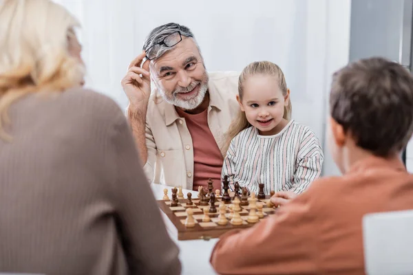 Hombre sonriente sosteniendo anteojos mientras juega al ajedrez con esposa y nietos - foto de stock