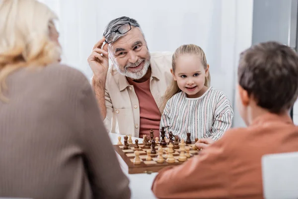 Smiling man holding eyeglasses while playing chess with wife and grandchildren — Stock Photo