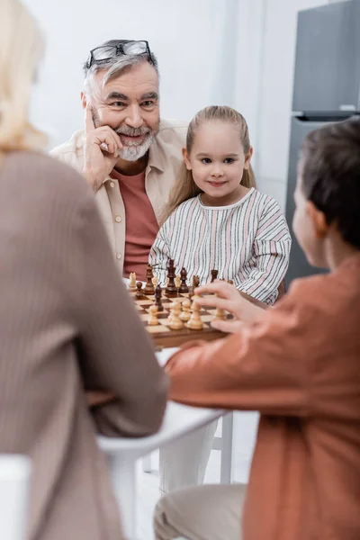 Joyeux homme âgé jouant aux échecs avec sa femme et ses petits-enfants au premier plan flou — Photo de stock