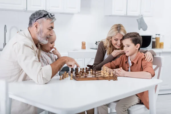 Senior man pointing at chessboard near grandchildren and wife in kitchen — Stock Photo