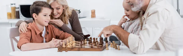 Hombre apuntando al tablero de ajedrez mientras juega ajedrez con nietos y esposa, estandarte - foto de stock