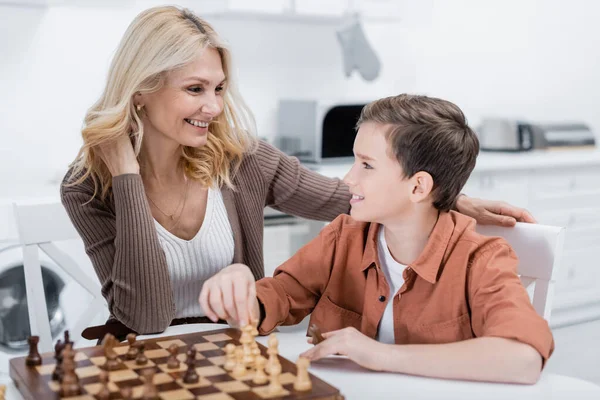 Sonriente chico mirando feliz abuelita mientras jugando ajedrez en cocina — Stock Photo