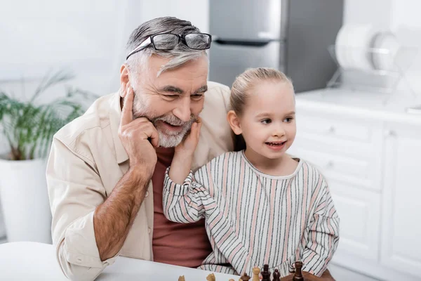 Smiling girl touching face of happy grandpa playing chess in kitchen — Stock Photo