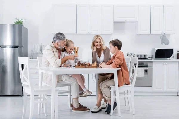 Hombre mayor señalando con el dedo mientras juega al ajedrez con esposa y nietos - foto de stock