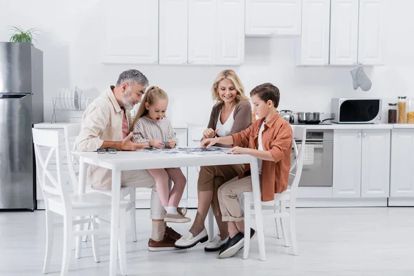 Feliz pareja jugando rompecabezas juego con nietos en la cocina - foto de stock