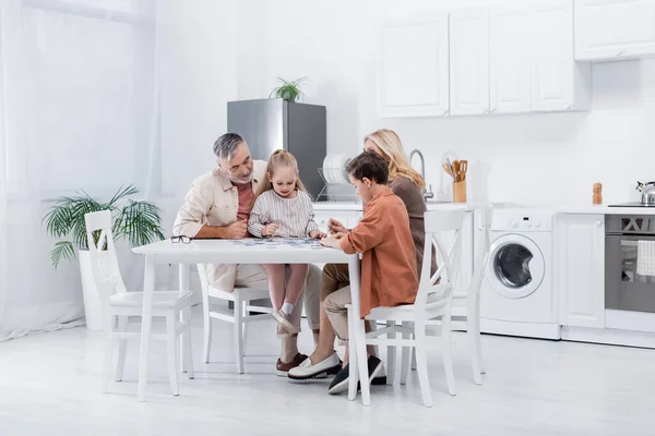 Abuelos con nietos jugando juego de rompecabezas en la cocina moderna - foto de stock