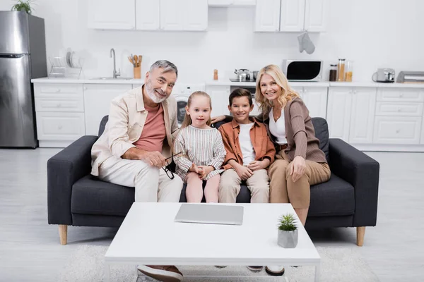 Heureux grands-parents avec des enfants regardant la caméra tout en étant assis sur le canapé près d'un ordinateur portable — Photo de stock