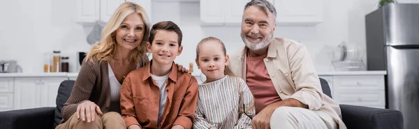 Счастливые дети сидят на диване с бабушкой и дедушкой и улыбаются в камеру, баннер — стоковое фото