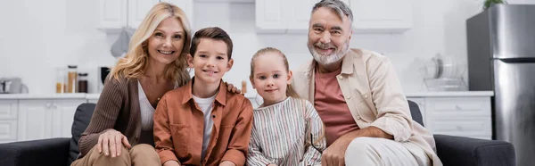 Crianças felizes sentadas no sofá com avós e sorrindo para a câmera, banner — Fotografia de Stock