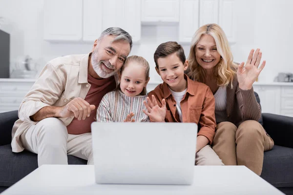 Alegre pareja con nietos agitando las manos durante el chat de vídeo en el ordenador portátil - foto de stock